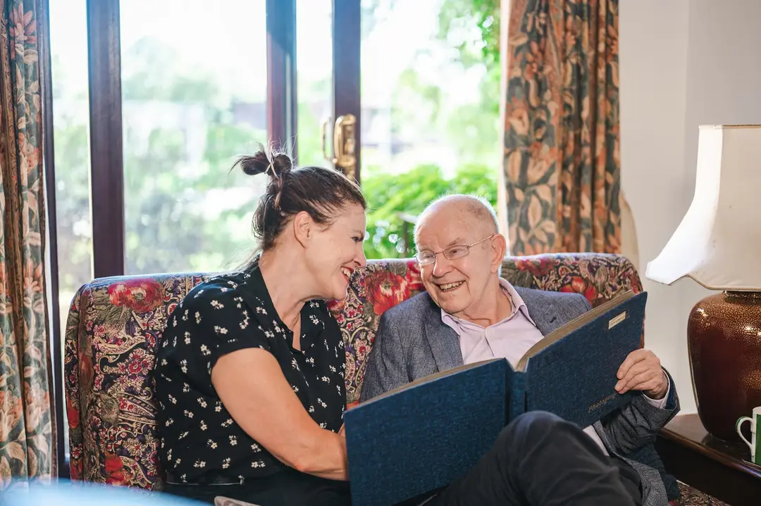 A care worker and an older man laughing over a photo album on a sofa