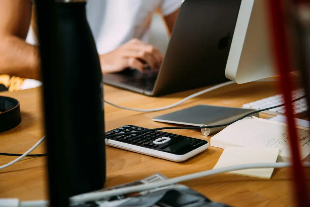 A desk with a calculator next to a laptop and a monitor