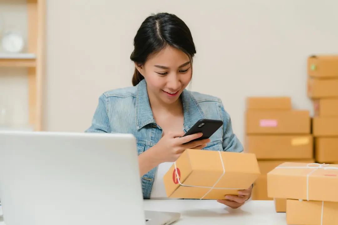 A woman checking a parcel on her phone beside a laptop and a stack of shipping boxes