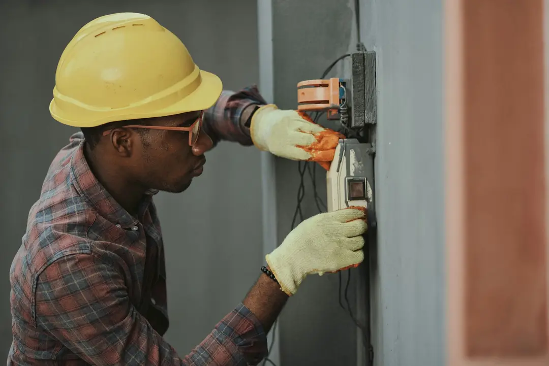 An electrician in a yellow hard hat and gloves wiring a wall box