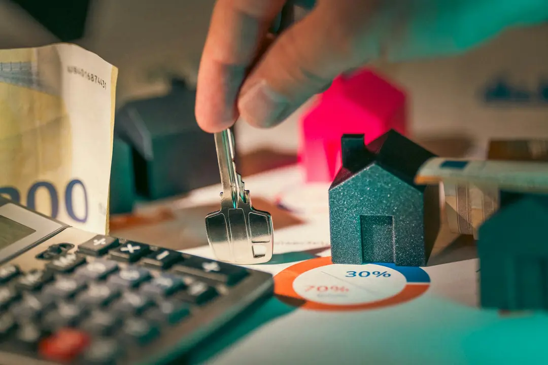 A hand holding a key above small model houses and a calculator