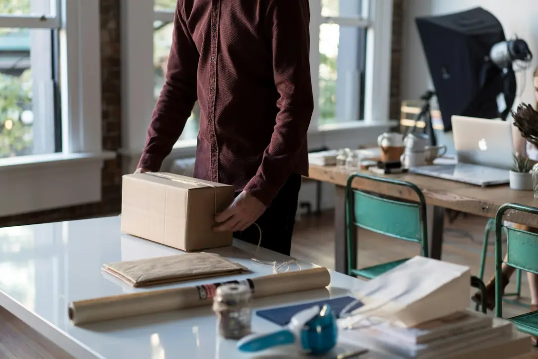 A person packing a cardboard box on a white table in a bright studio with a laptop and lighting gear