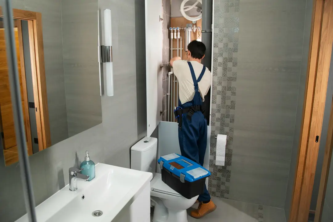 A plumber in overalls fitting pipes behind a bathroom wall